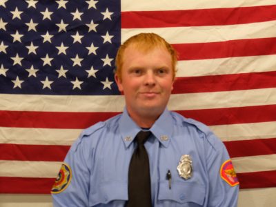 A person with red hair is wearing a blue uniform with badges and a black tie, standing in front of an American flag.