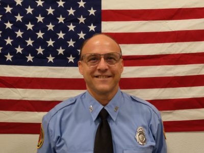 A man in a blue uniform with a badge and black tie stands in front of an American flag, smiling. He is wearing glasses and a patch is visible on his sleeve.