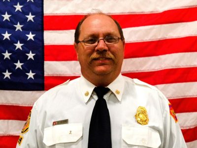 A middle-aged man in a white firefighter uniform with badges, smiling in front of an american flag.