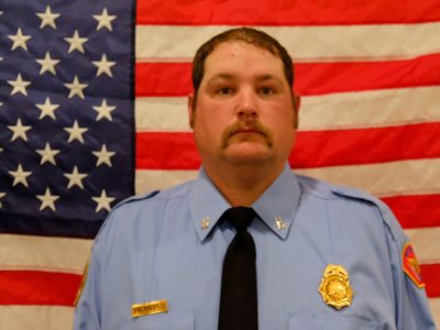 A portrait of Captain Andrew Venables in blue uniform, with a badge and name tag, standing in front of an american flag.