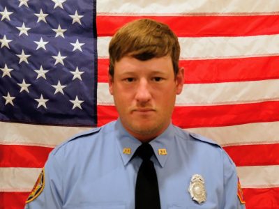A police officer in a blue uniform, featuring a badge and patches, poses solemnly in front of an american flag.