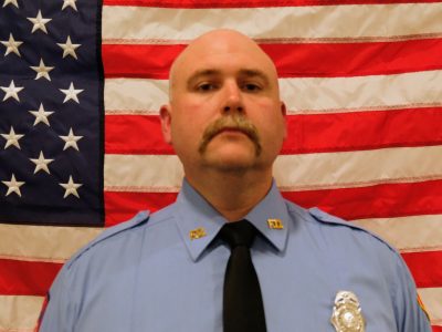 Darreck Kenjura Lieutentant with a serious expression, wearing a blue uniform with badges, stands in front of an american flag.