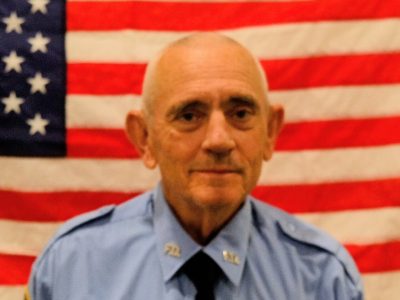 An elderly male police officer in uniform, with a serious expression, sitting in front of an american flag.