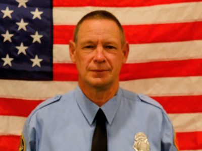 Firefighter Alan Winkelmannwith a badge and tie, stands in front of an american flag. he has a serious expression.
