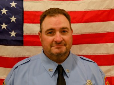 A portrait of a caucasian male firefighter in a blue uniform with badges, smiling slightly in front of an american flag.