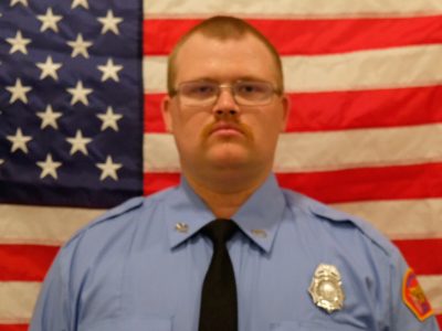 Jacob Tieman a uniformed firefighter stands solemnly in front of an american flag, wearing glasses and a serious expression. his badge is visible on his light blue shirt.