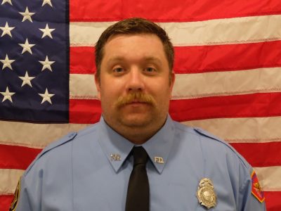 Firefighter Matt Martin in a blue firefighter uniform with badges, standing in front of an american flag. he has a mustache and appears serious.