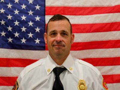 A man in a white uniform with a badge, smiling slightly, sits in front of an american flag, displaying a professional demeanor.