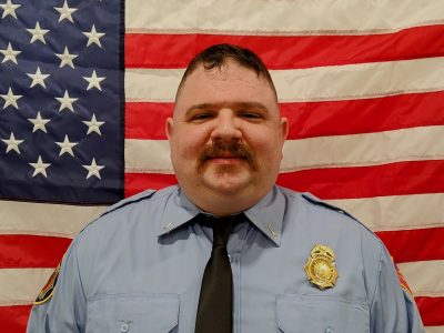 Lietenant Daniel Moehlmann in a blue uniform with a badge and tie, smiling in front of an american flag.