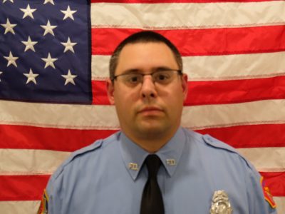 Matt Metzger Firefighter in a blue uniform with a badge, standing in front of an american flag, looking directly at the camera. the focus is slightly blurred.