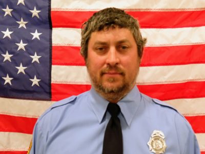 Rallin Wellmann, Firefighter in a blue uniform and badge stands in front of an american flag. he has a slight smile and is wearing a winter hat.